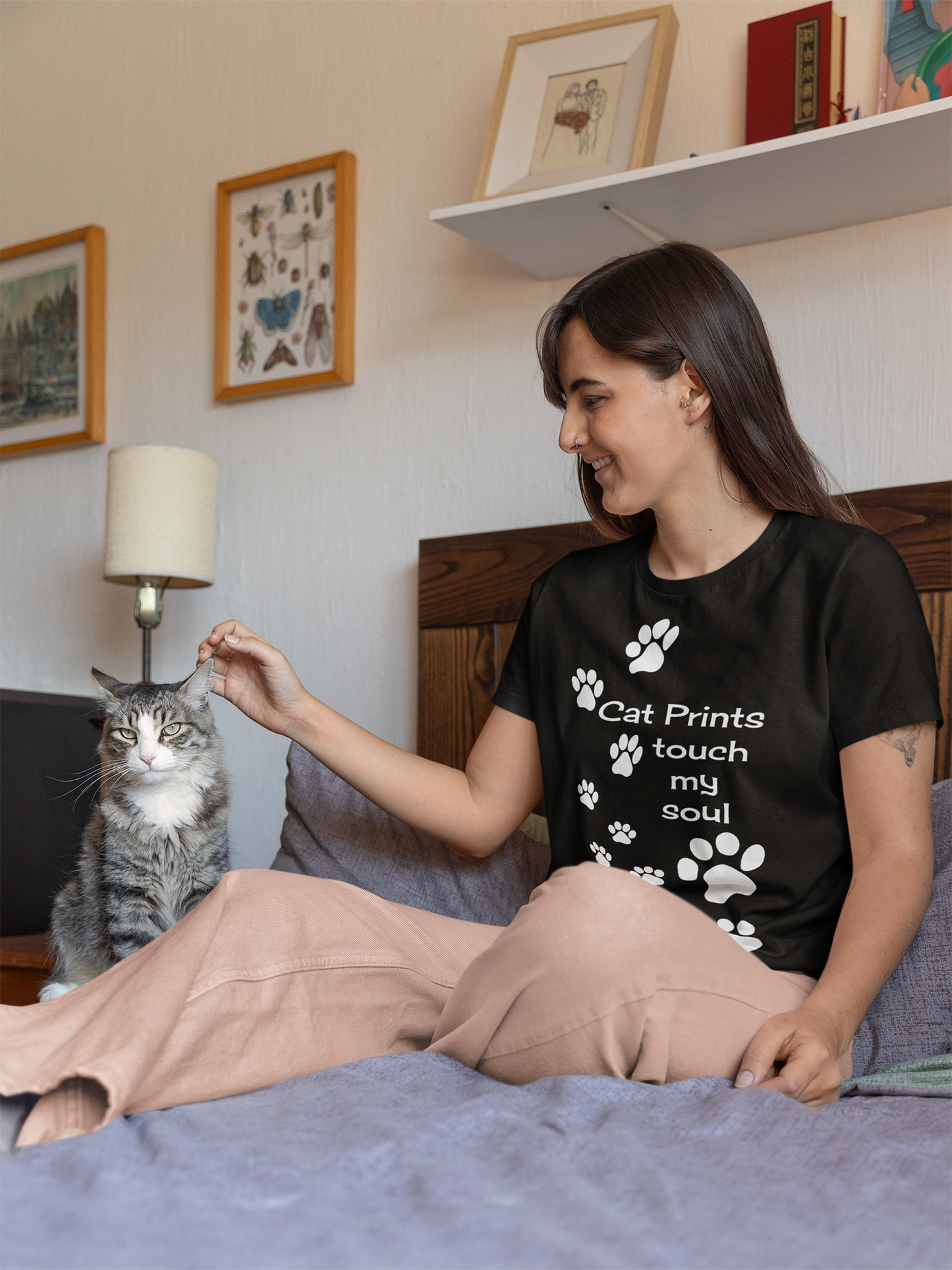 Frau sitzt mit grauer Katze auf dem Bett. Sie trägt eine beige Hose und ein schwarzes T-Shirt mit Cat Prints und Spruch "Cat Prints touch my soul". Hinten an der Wand hängen Bilder.