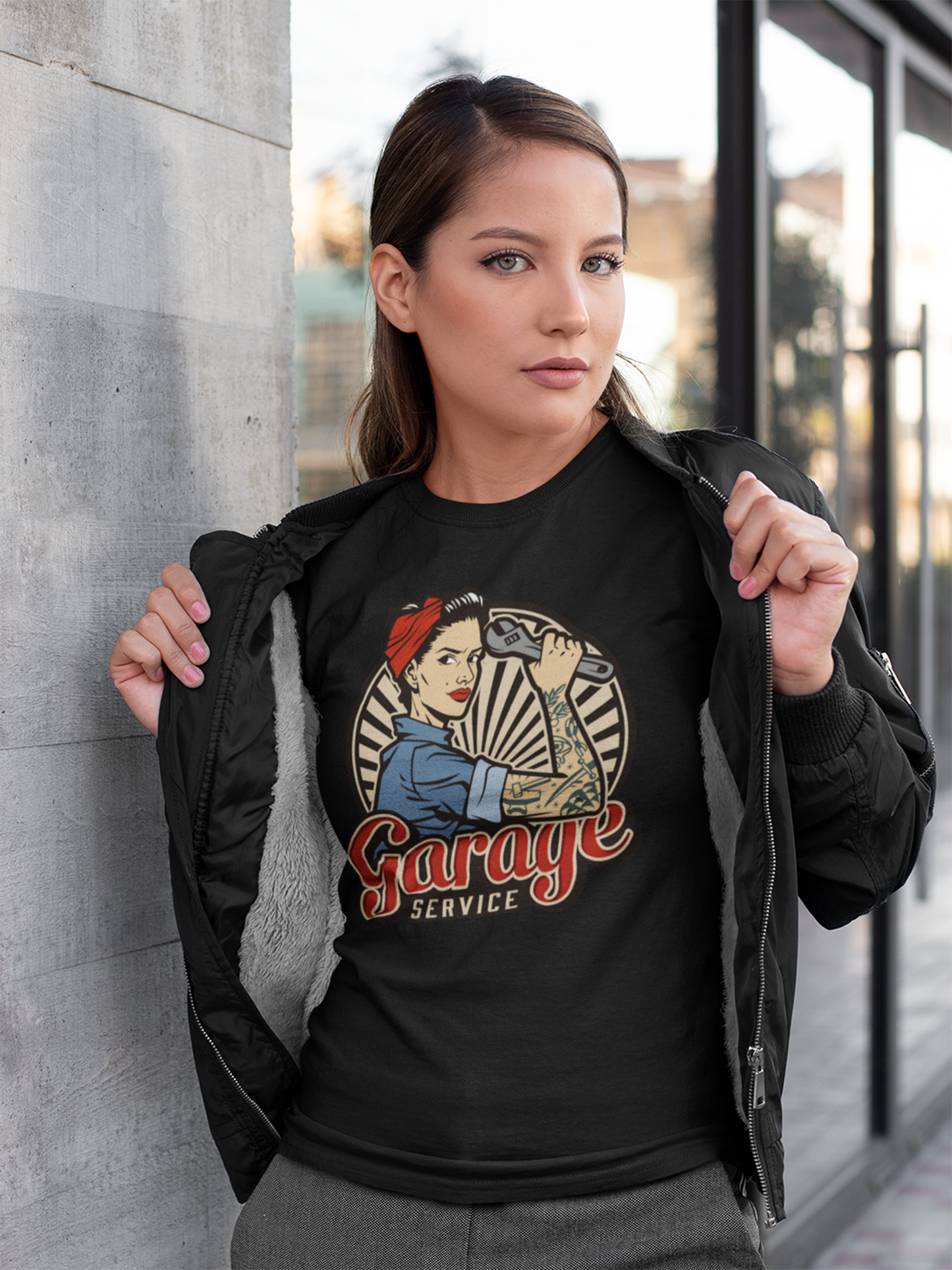 Junge Frau trägt eine schwarze offene Jacke und darunter ein schwarzes T-Shirt mit Rockabilly-Motiv einer starken Frau mit Werkzeug, Oberkörperansicht
