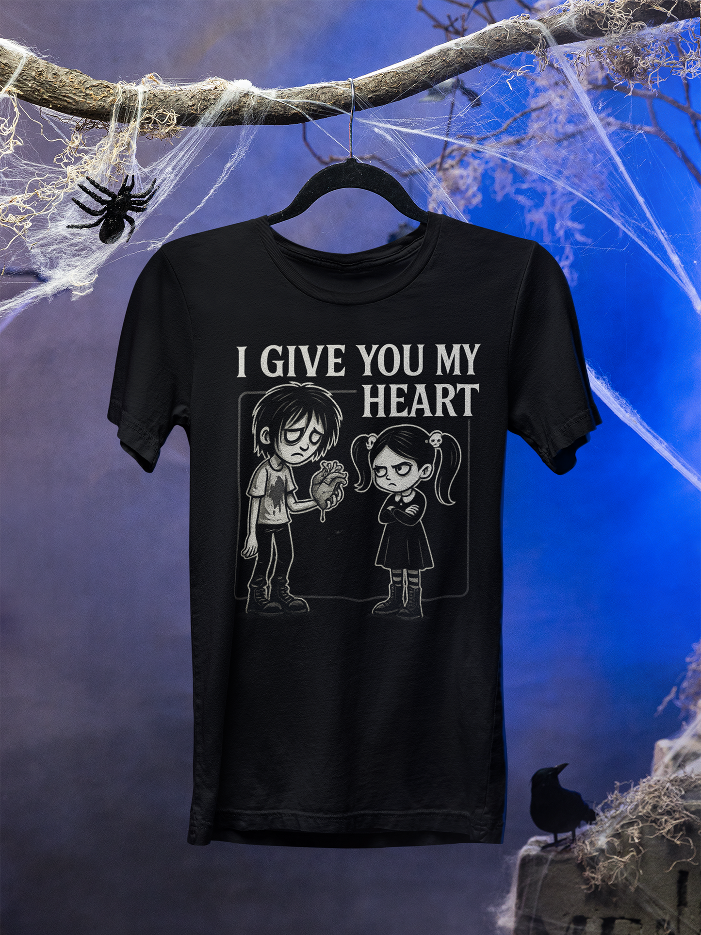 Schwarzes T-Shirt mit Gothic Junge, Herz in der Hand, Gothic Mädchen mit Skull-Haarspangen, Schriftzug „I Give You My Heart“. Hintergrund blau mit Spinnenweben und Halloween-Deko.