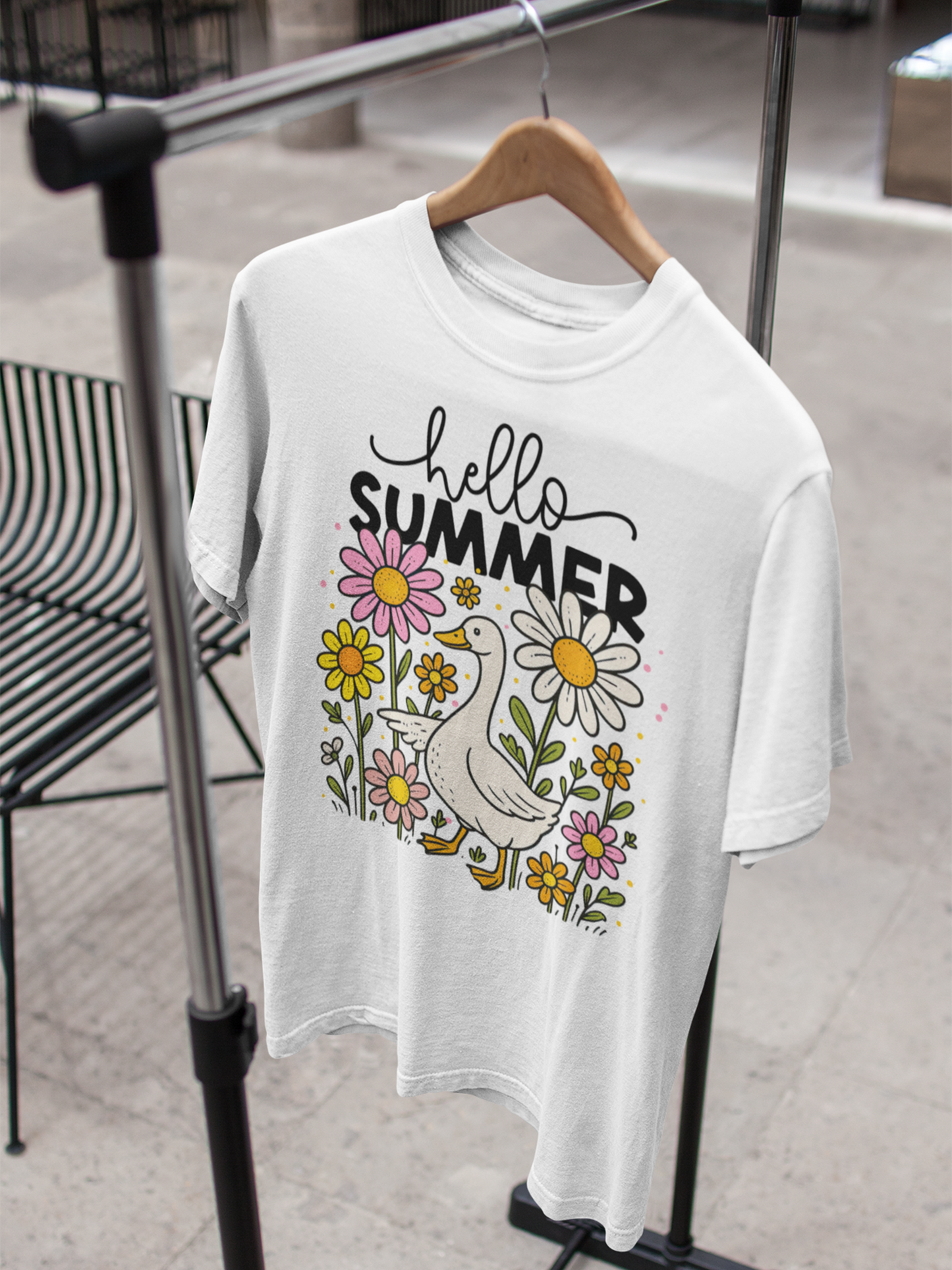 Weißes T-Shirt mit Gans und Blumenmotiv sowie Schriftzug „Hello Summer“, hängend auf einem Holzkleiderbügel am Kleiderständer