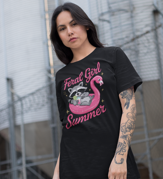 Mädchen trägt schwarzes T-Shirt mit Waschbär auf pinkem Flamingo-Schwimmreifen, Cocktail in der Pfote und Schriftzug „Feral Girl Summer“