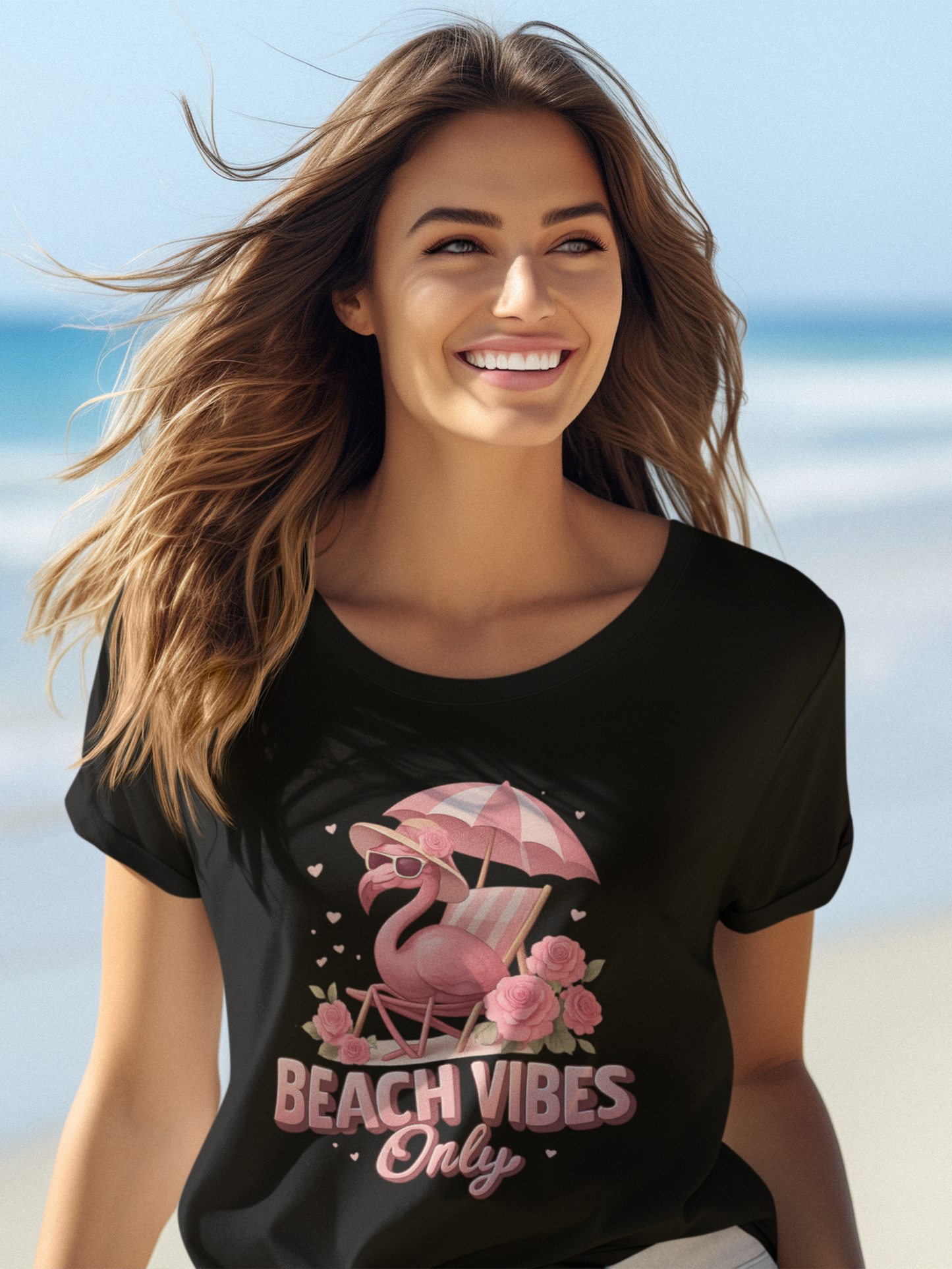 Langhaarige, lachende Frau trägt ein schwarzes Shirt mit Flamingo-Motiv und der Aufschrift "Beach Vibes Only", im Hintergrund verschwommen das Meer.