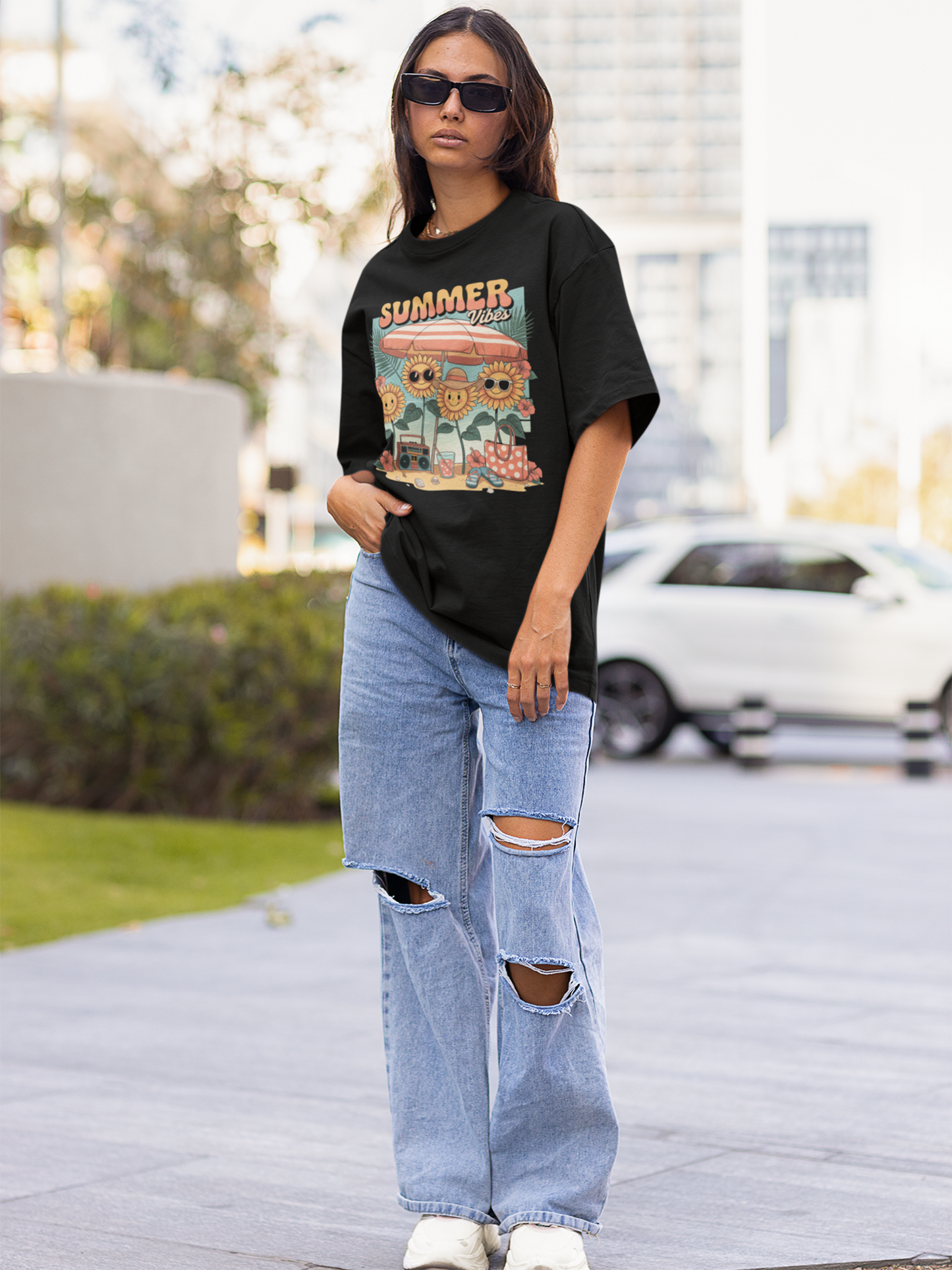 Frau mit Sonnenbrille steht auf der Straße. Sie hat eine Jeanshose und ein schwarzes Shirt mit bunten Print an.
