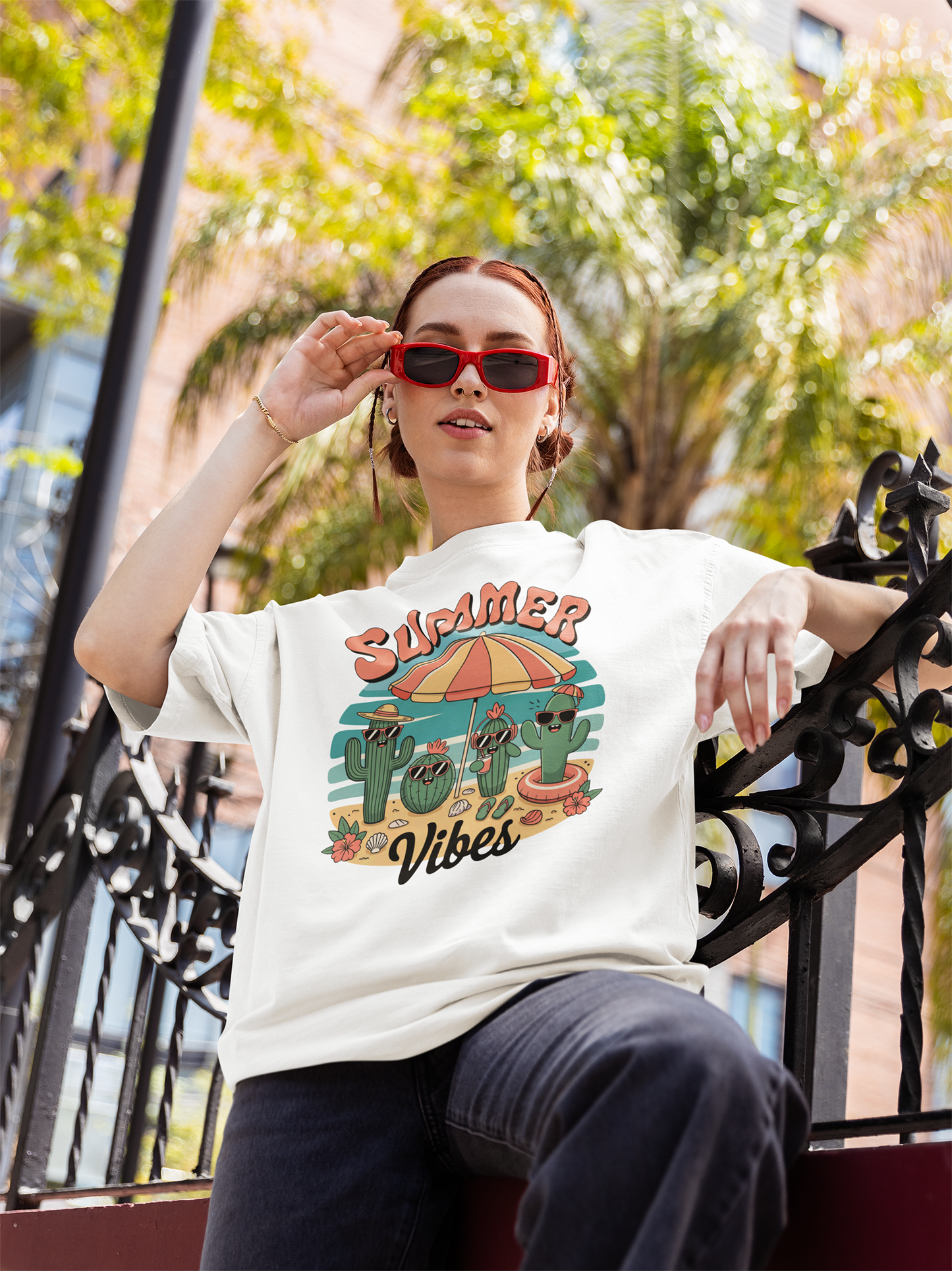 Junge Frau mit Sonnenbrille lehnt am Zaun und trägt ein weißes Shirt mit Kaktus-Print und Aufdruck "Summer Vibes"