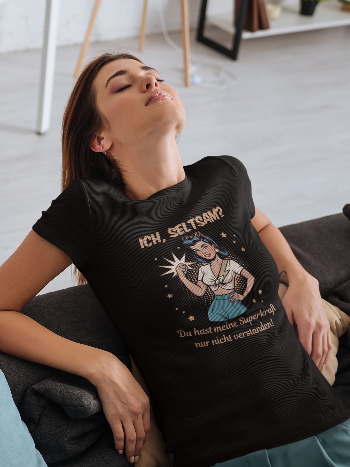 Junge Frau auf Sofa trägt schwarzes T-Shirt mit Motiv: Retro-Girl mit Sonnenbrille im 50s-Stil, mit Text „Ich, seltsam? Du hast nur meine Superkraft nicht verstanden!“