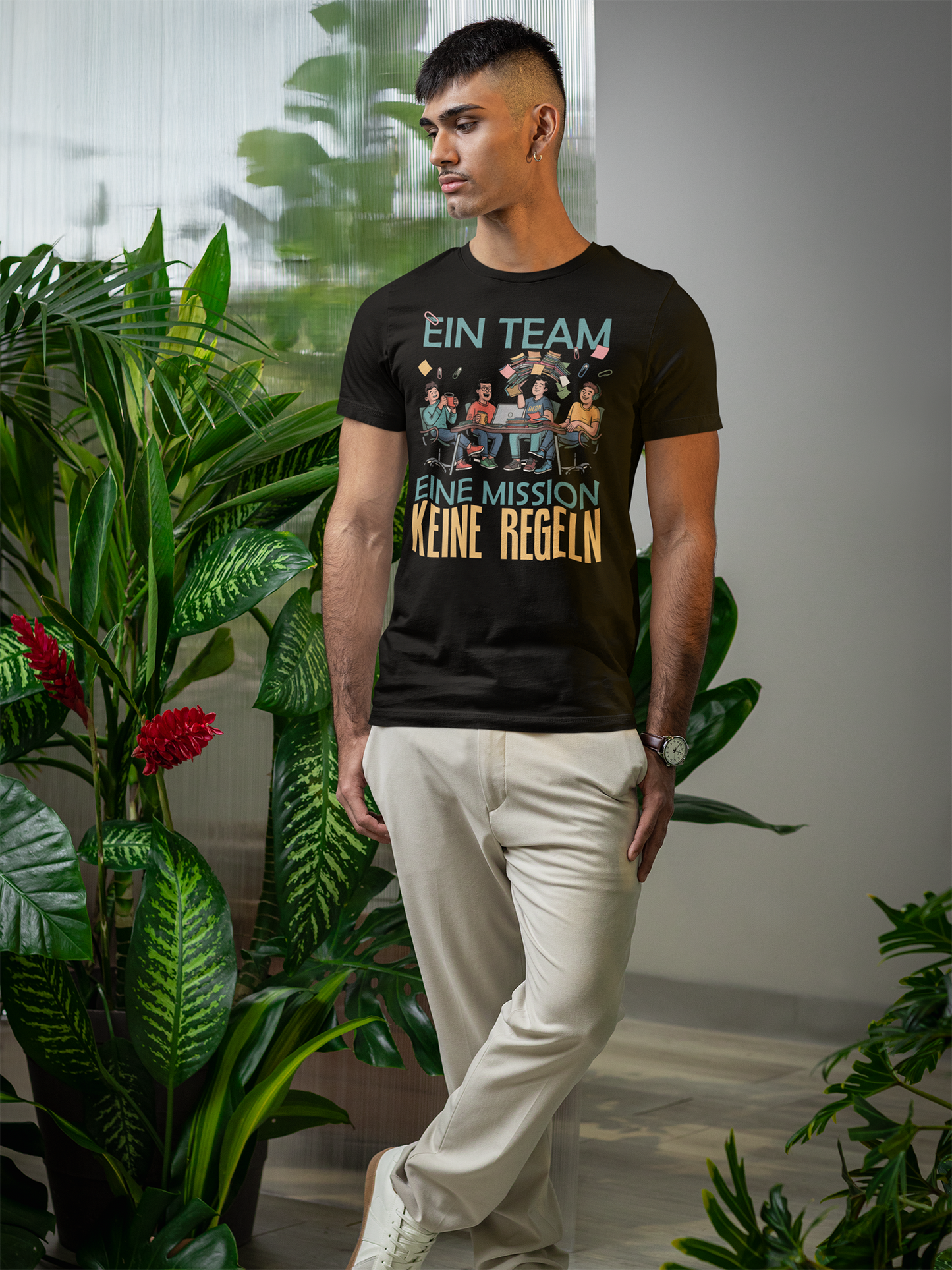Junger Mann, stehend, vor grünen Pflanzen. Er trägt eine weiße Hose und ein weißes Shirt mit Print: "Ein Team. Eine Mission. Keine Regeln"