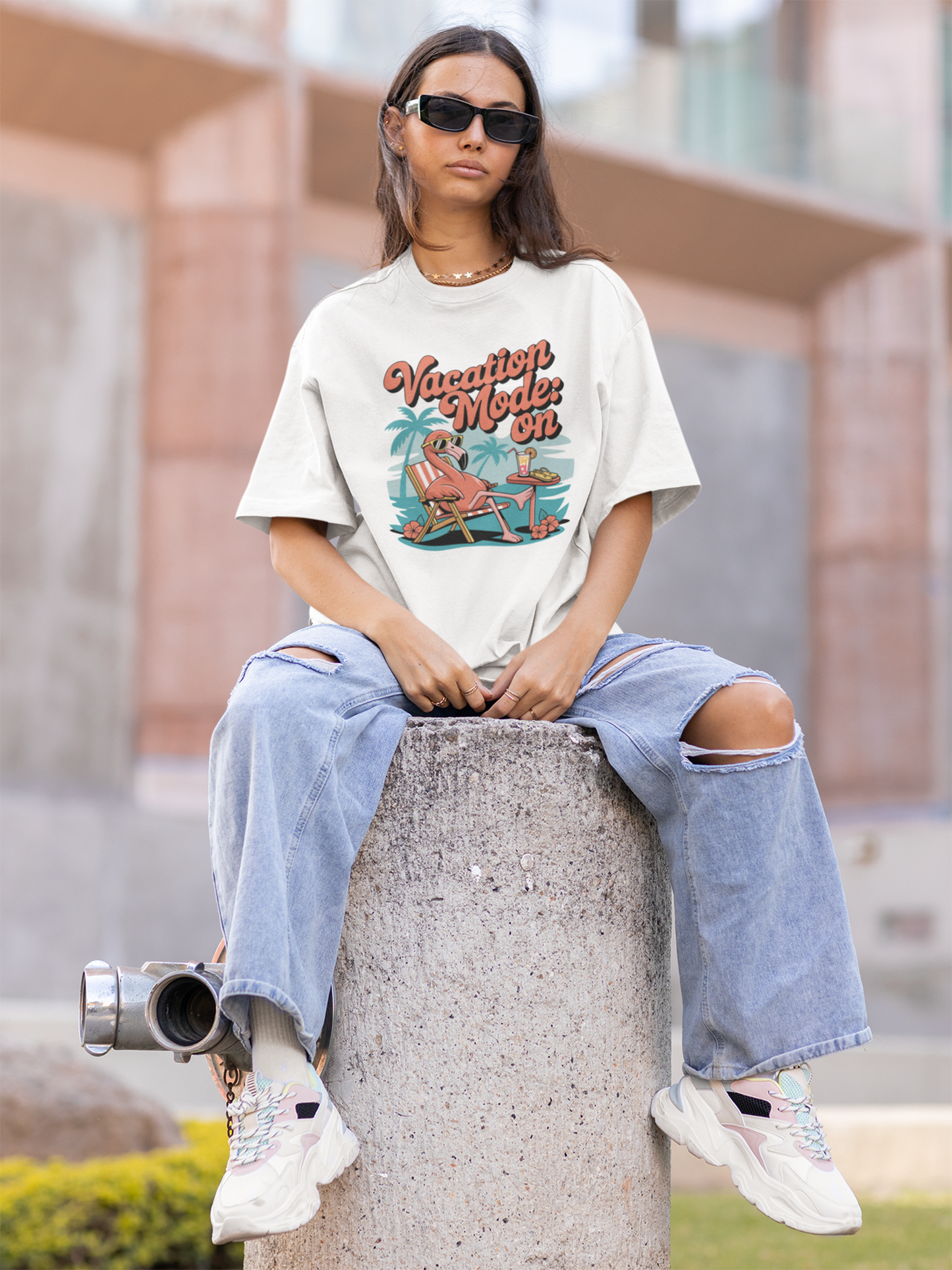 Junge Frau, sitzend auf einer Betonsäule, in kompletter Ansicht trägt ein weißes T-Shirt mit chillendem Flamingo und dem Text „Vacation Mode: On“