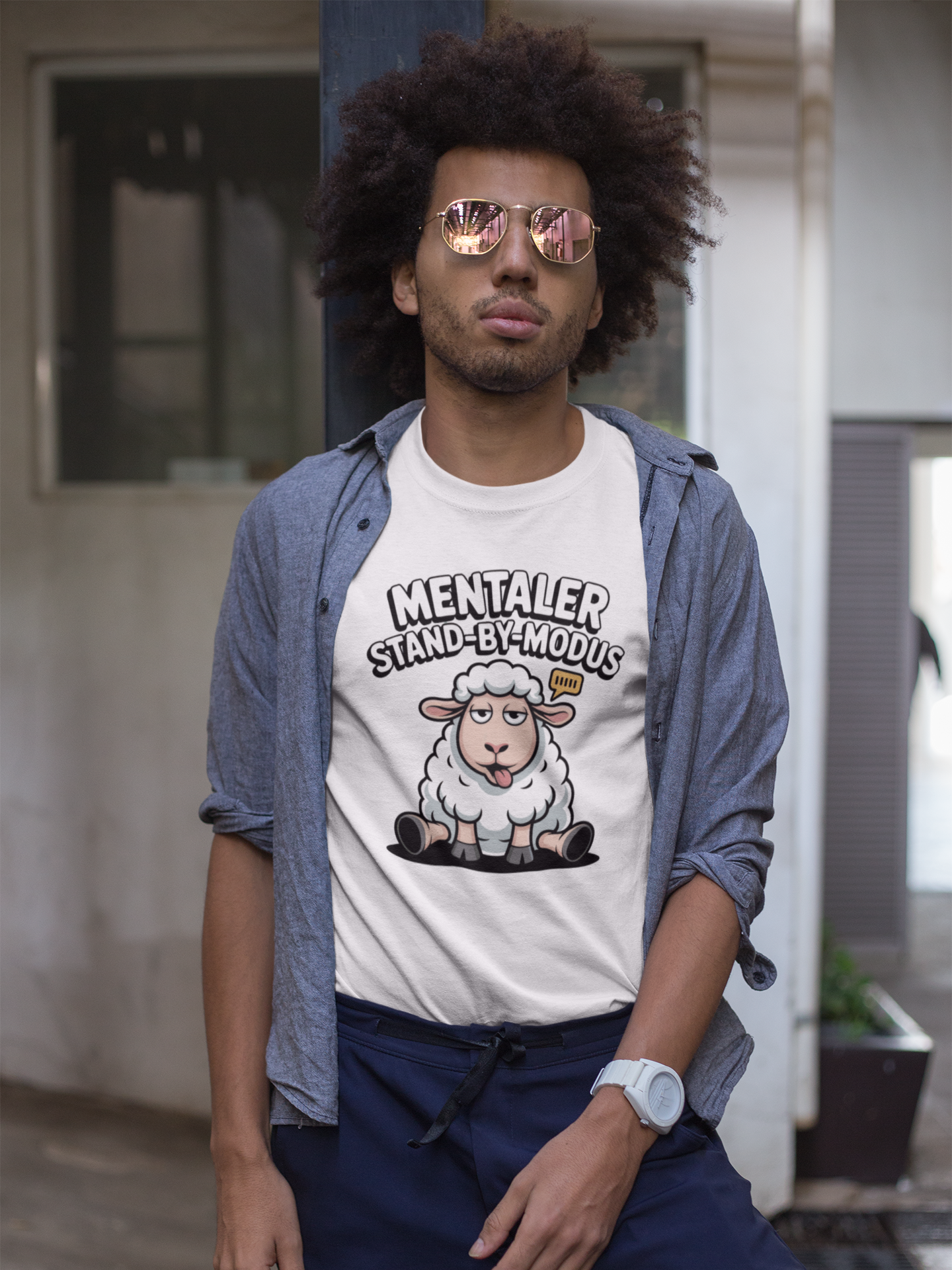Junger schwarzer Mann mit offenem Jeans-Hemd trägt darunter weißes T-Shirt mit müdem Schaf und Text „Mentaler Stand-By-Modus“ (Oberkörperaufnahme)