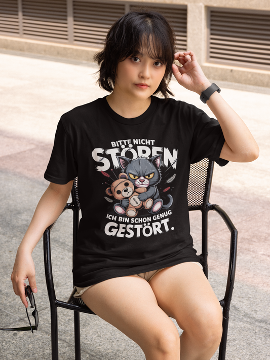 Frau, auf Stuhl sitzend, mit kurzer Hose, trägt schwarzes T-Shirt mit dem Motiv „Bitte nicht stören. Ich bin schon genug gestört.“ mit Voodoo-Katze (Oberkörper)