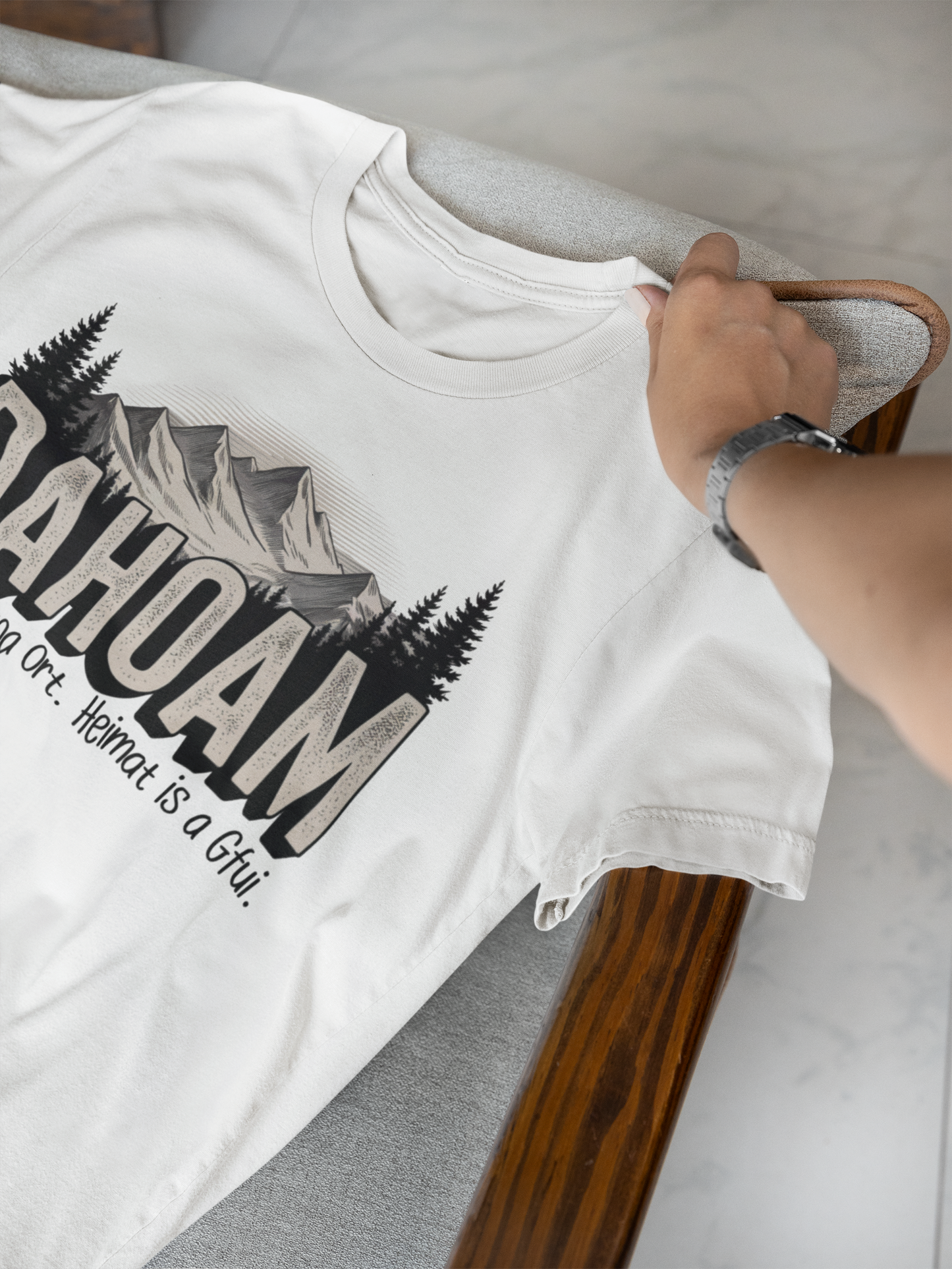 Weißes T-Shirt, liegend, Teilansicht mit Hand, mit dem Spruch „Dahoam. Heimat is koa Ort. Heimat is a Gfui“ und Bergmotiv