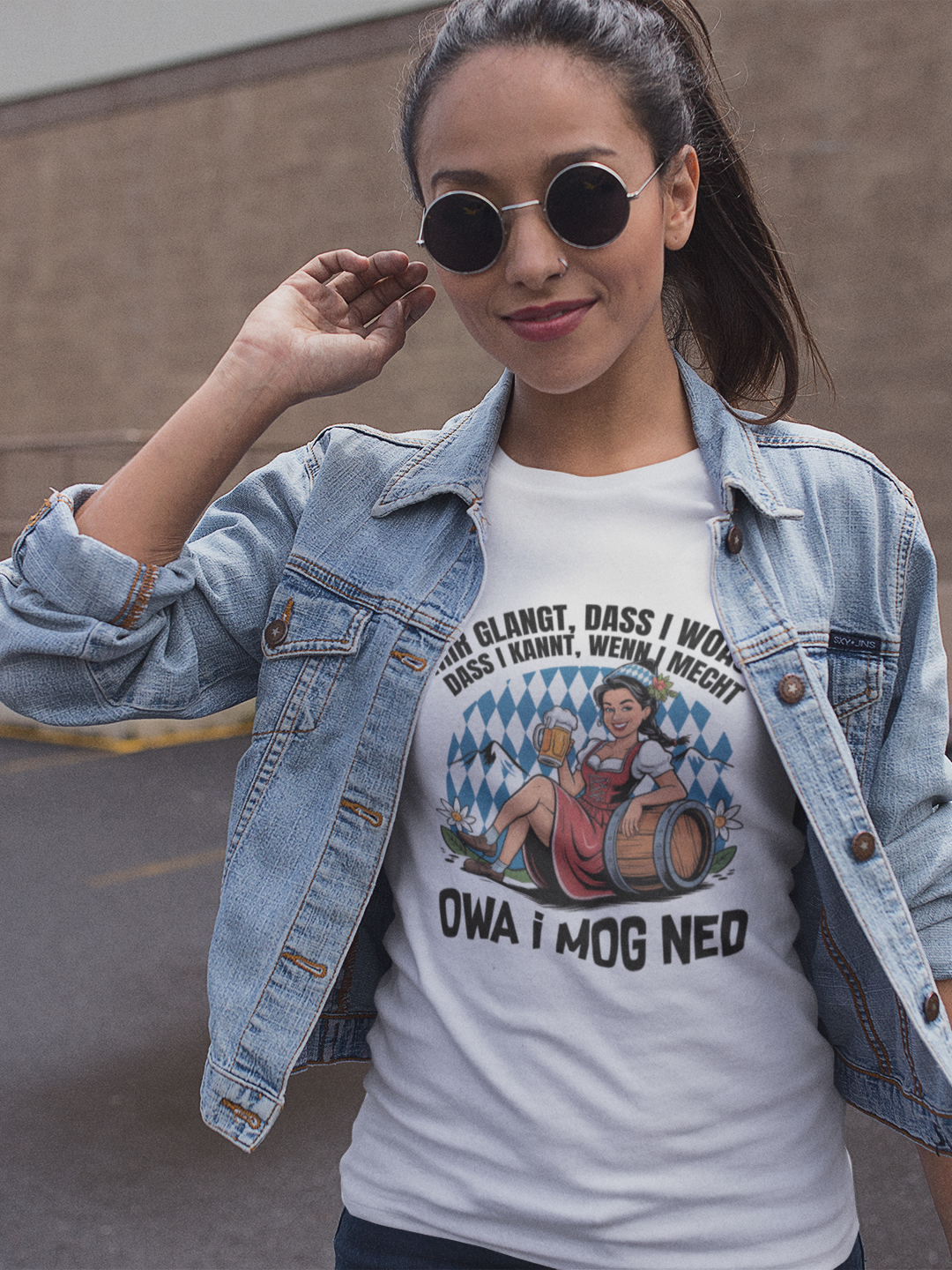 Frau mit Sonnenbrille und offener Jenasjacke, darunter trägt sie ein weißes T-Shirt mit Motiv „Owa i mog ned“ und bayrischem Madl Motiv(Oberkörper)