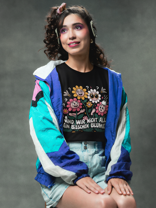 Junge Frau, auf Stuhl sitzend, 80er Jahre Look, offene blaue Jacke, darunter trägt sie ein schwarzes T-Shirt mit buntem Blumenmotiv und Spruch „Sind wir nicht alle ein bisschen Blume?“ (Oberkörper)