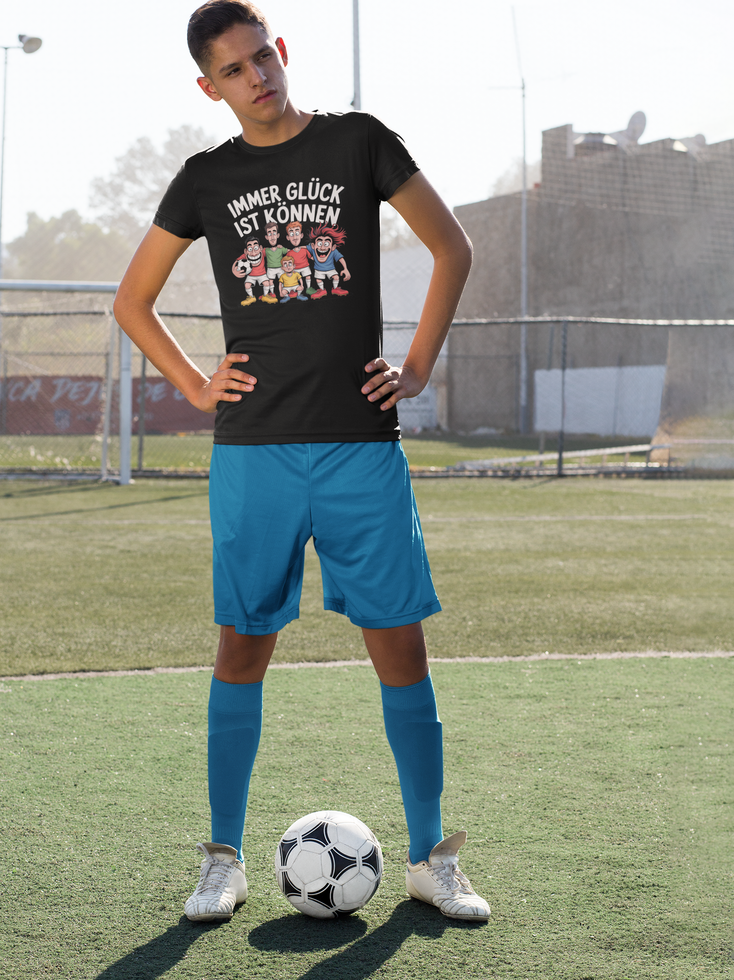 Fußballspieler auf rasen mit Ball, blaue Hose und schwarzes Shirt mit Spruch „Immer Glück ist Können“ und Fußballteam-Motiv (Ganzkörper)