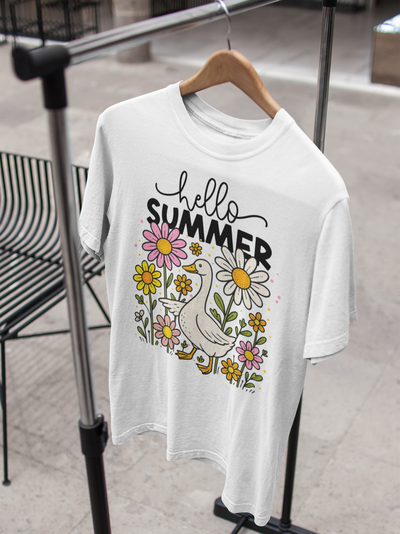 Weißes T-Shirt mit Gans und Blumenmotiv sowie Schriftzug „Hello Summer“, hängend auf einem Holzkleiderbügel am Kleiderständer