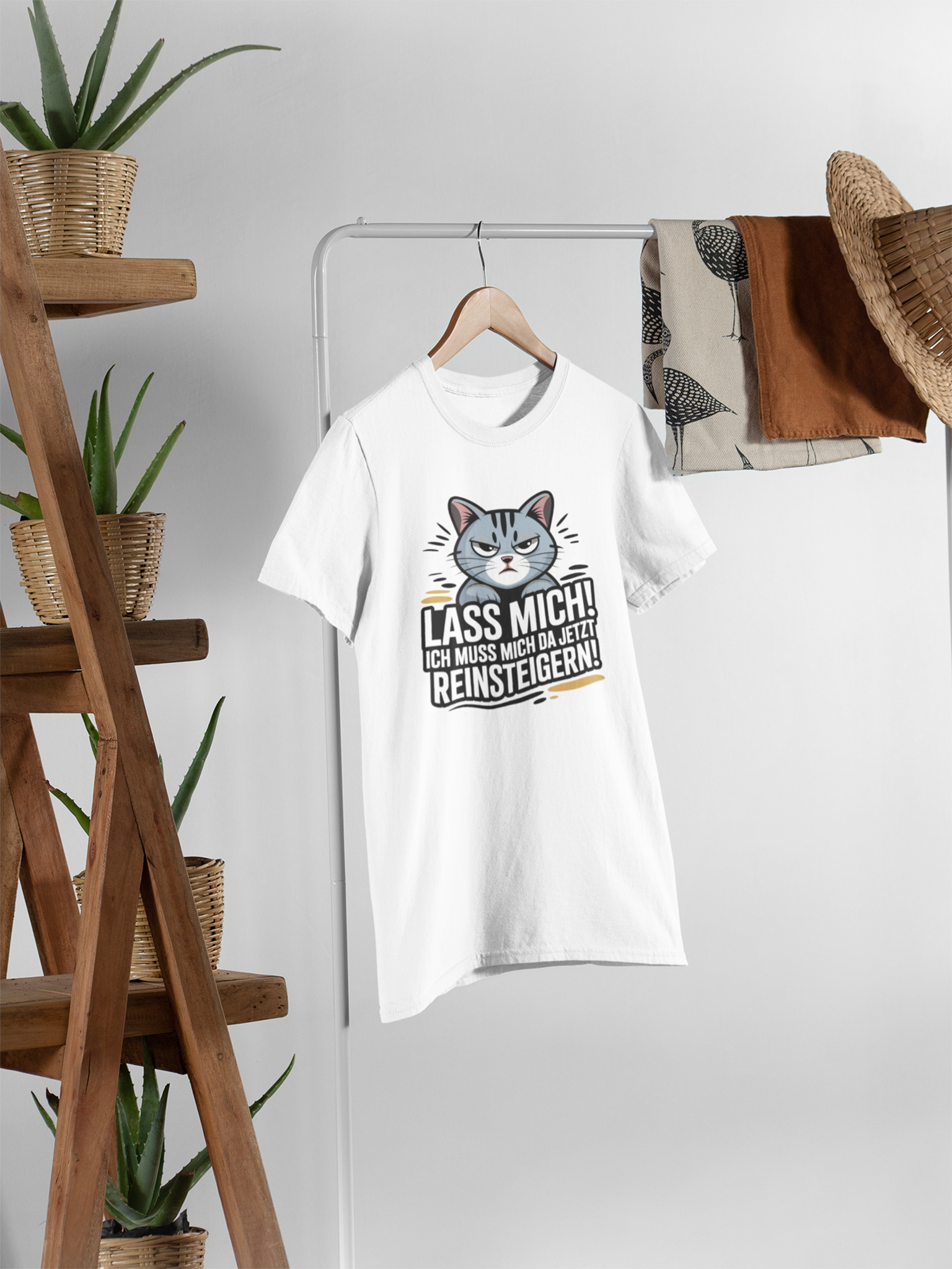 Mittig ein Kleiderständer mit weißen Shirt mit großer Katze und Spruch "Lass mich! Ich muss mich da jetzt reinsteigern!". Links im Bild eine Holztreppe mit Pflanzen.