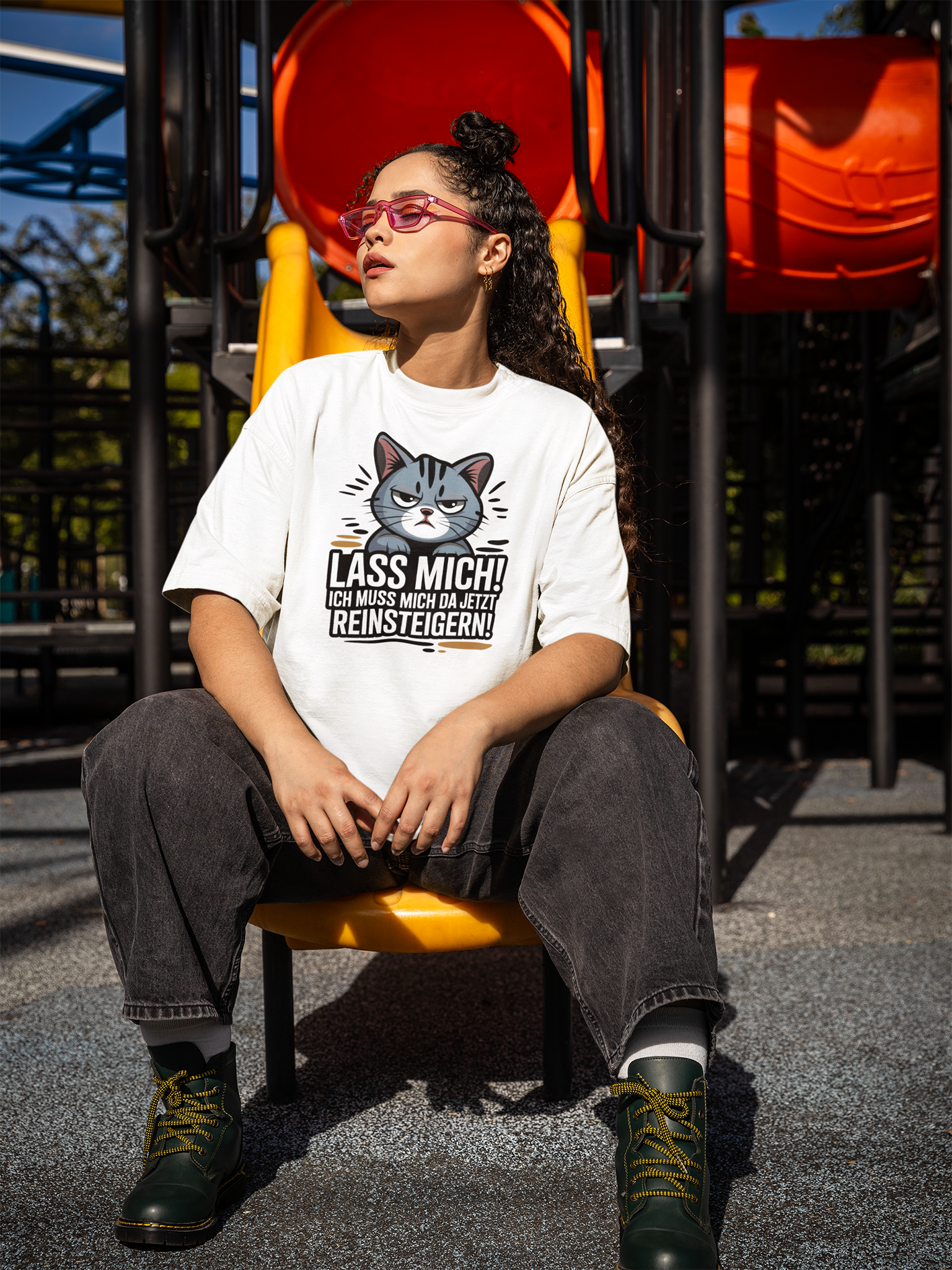 Junge Frau mit Brille sitzt auf gelber Rusche. Sie trägt eine graue Jeanshose und ein weißes Shirt mit großem Katzen-Motiv und den Spruch:  "Lass mich! Ich muss mich da jetzt reinsteigern!"