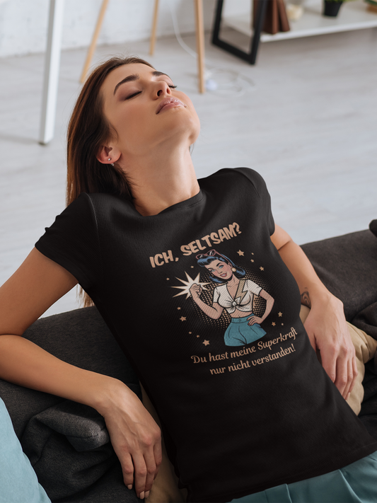 Junge Frau auf Sofa trägt schwarzes T-Shirt mit Motiv: Retro-Girl mit Sonnenbrille im 50s-Stil, mit Text „Ich, seltsam? Du hast nur meine Superkraft nicht verstanden!“