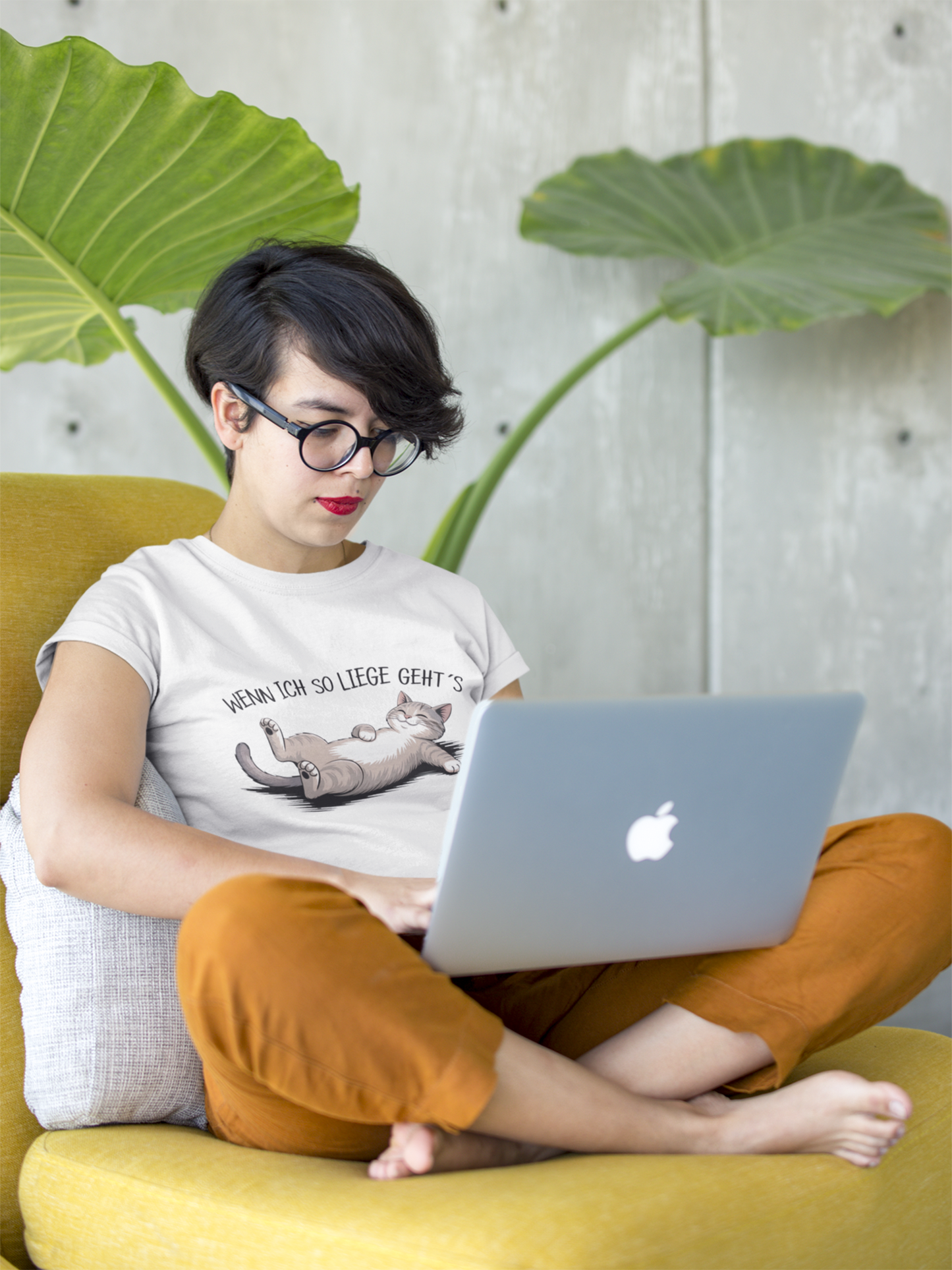 Junge Frau sitzt im Schneidersitz mit Laptop auf gelben Sofa. Sie trägt ein weißes Sommer-T-Shirt mit Katze & Spruch, Ganzkörperansicht