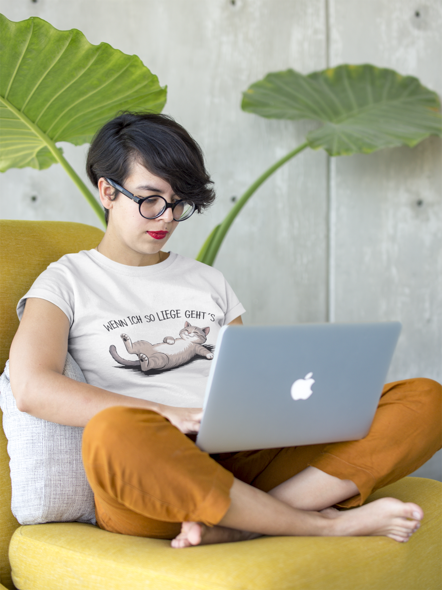 Junge Frau sitzt im Schneidersitz mit Laptop auf gelben Sofa. Sie trägt ein weißes Sommer-T-Shirt mit Katze & Spruch, Ganzkörperansicht