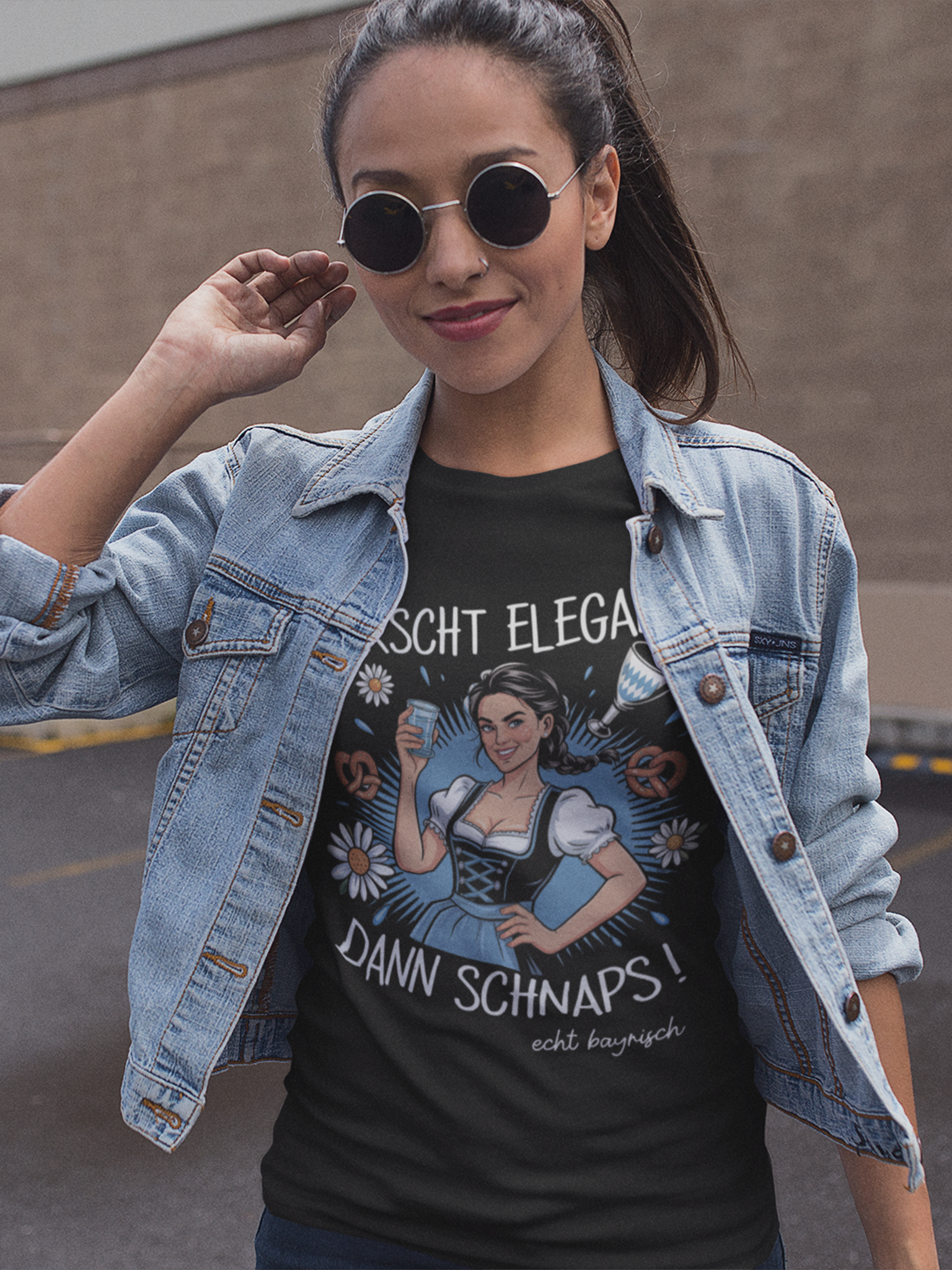 Junge Frau mit Jeansjacke und Sonnenbrille. Sie trägt schwarzes T-Shirt mit Dirndl-Motiv und Spruch „Erscht elegant dann Schnaps“, Oberkörperansicht.