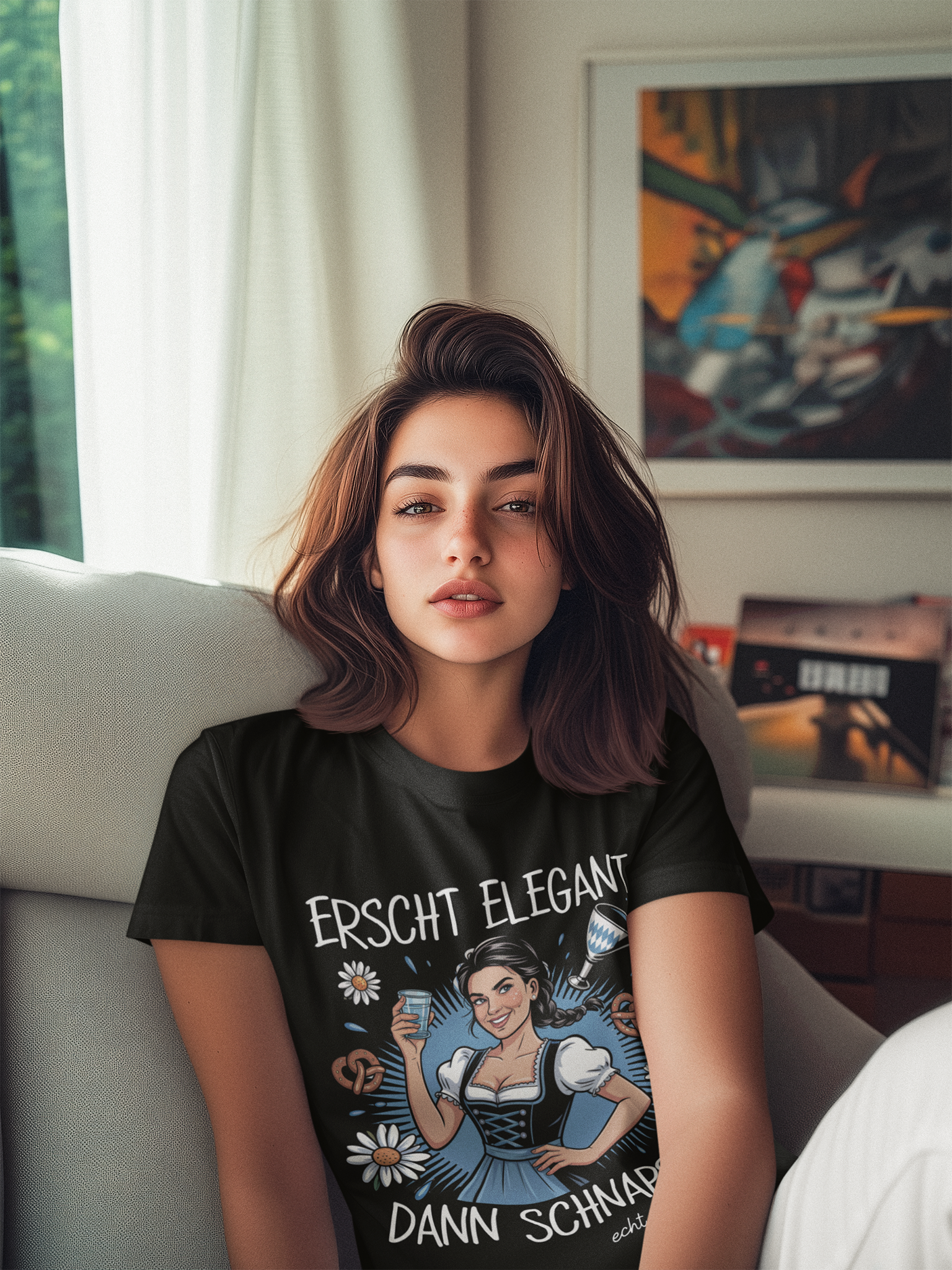 Frau sitzt auf Sofa, sie trägt schwarzes T-Shirt mit Dirndl-Motiv und Spruch „Erscht elegant dann Schnaps“, Oberkörperansicht