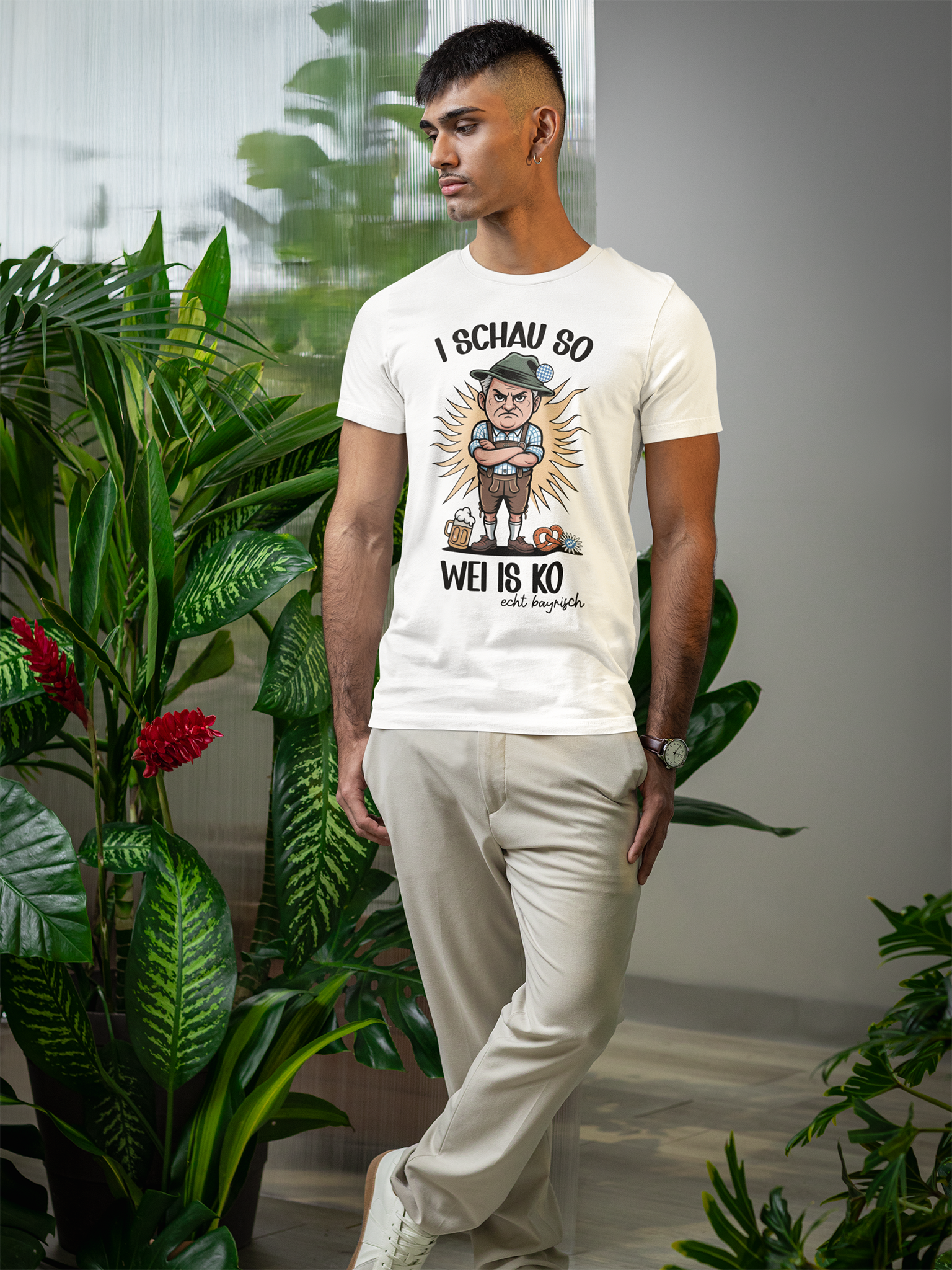 Mann trägt weißes T-Shirt mit Trachtenmotiv und bayerischem Spruch, Ganzkörperansicht. Im Huntergrund grüne Pflanzen.
