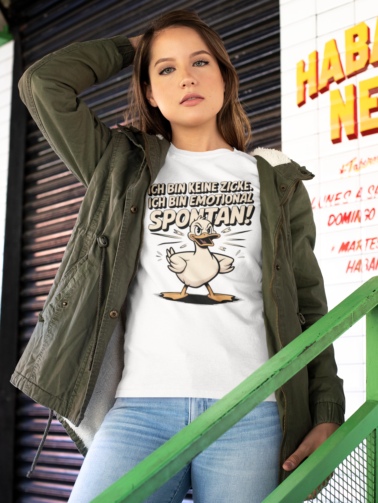 Junge Frau mit offener grüner Jacke, darunter trägt sie ein weißes T-Shirt mit Ente und Spruch „Ich bin keine Zicke, ich bin emotional spontan“ (Oberkörperaufnahme)