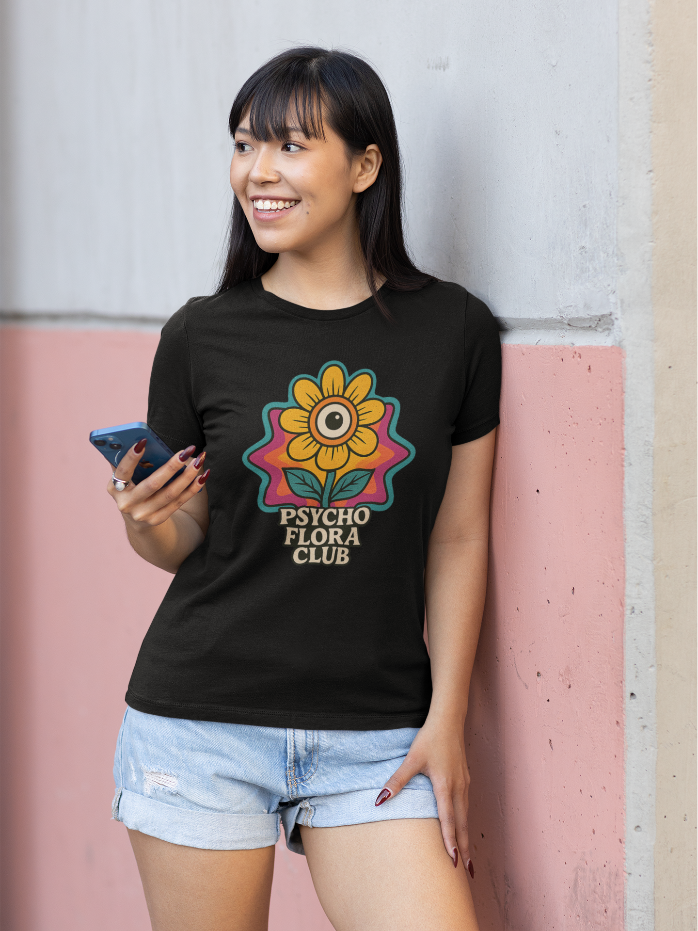 Junge Frau, lachend, mit Handy in der Hand, lehnt an Wand. Sie trägt schwarzes Shirt mit einzelner Blume und „Psycho Flora Club“-Schriftzug (Oberkörper)