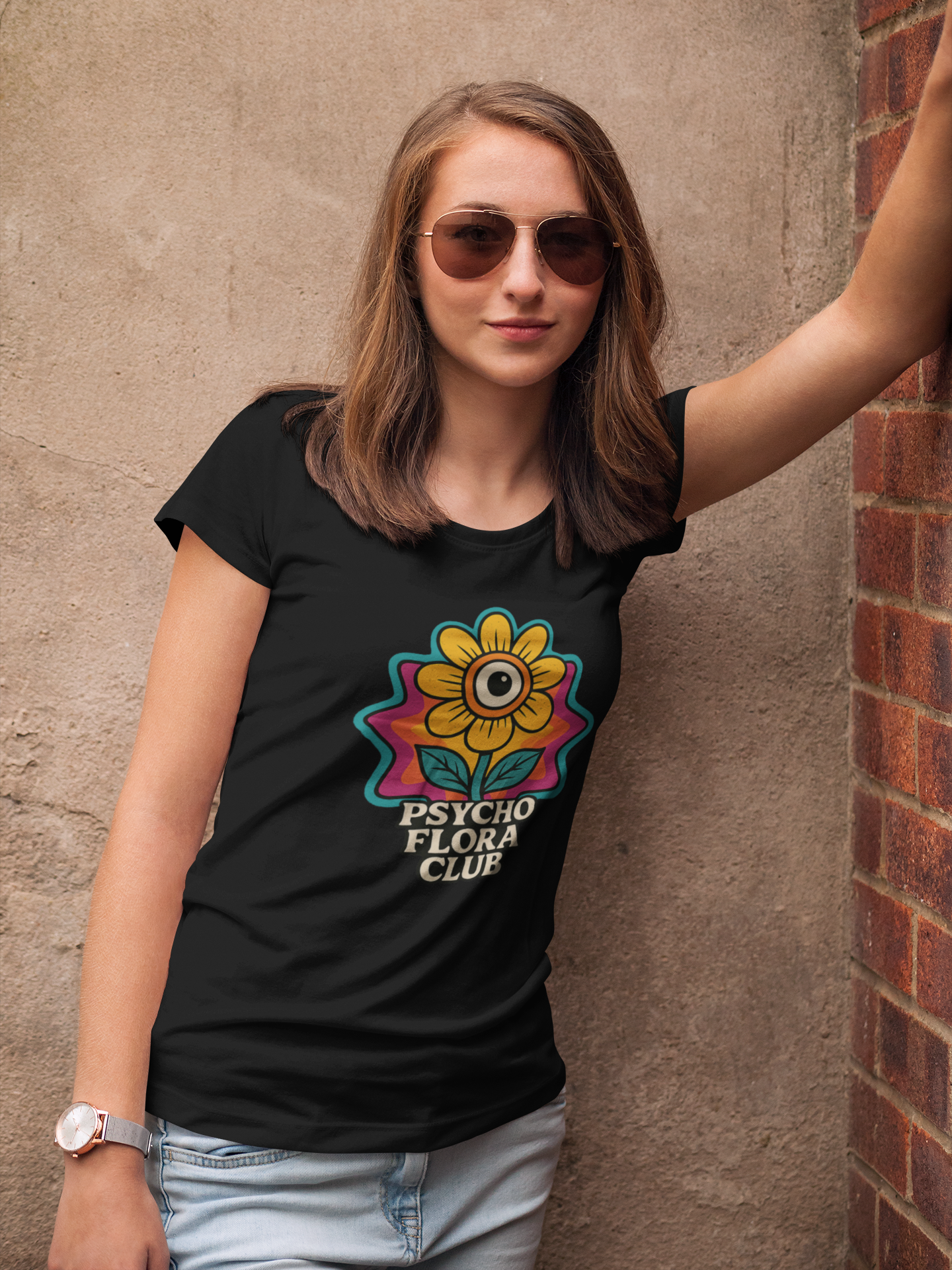 Junge Frau mit Sonnenbrille, lehnt an der Mauer, sie trägt schwarzes Shirt mit einzelner Blume und „Psycho Flora Club“-Schriftzug (Oberkörper)