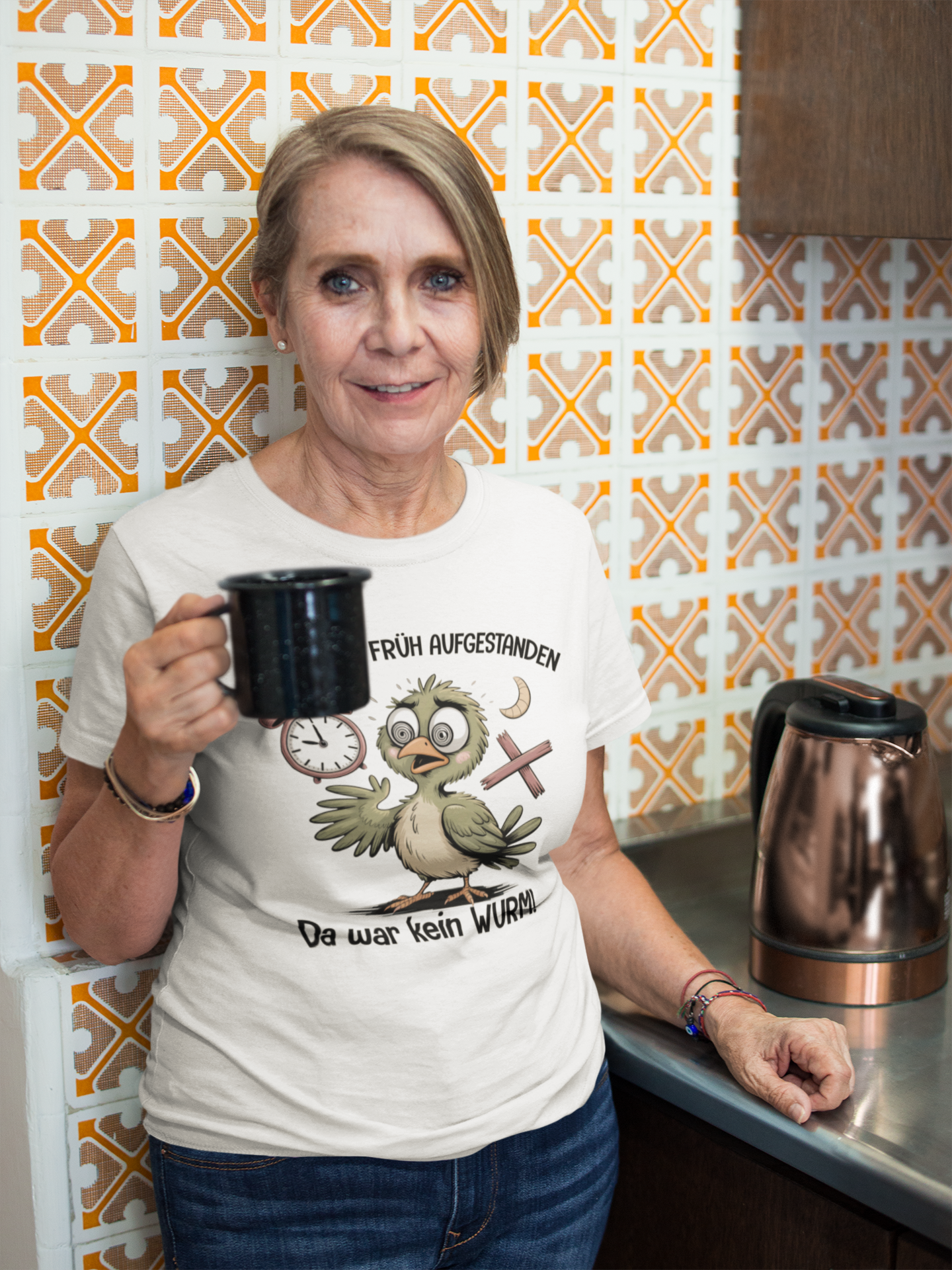 Ältere Frau in der Küche mit schwarzer Kaffeetasse. Sie trägt ein weißes T-Shirt mit Vogel-Motiv und Text „Bin extra früh aufgestanden – Da war kein Wurm“