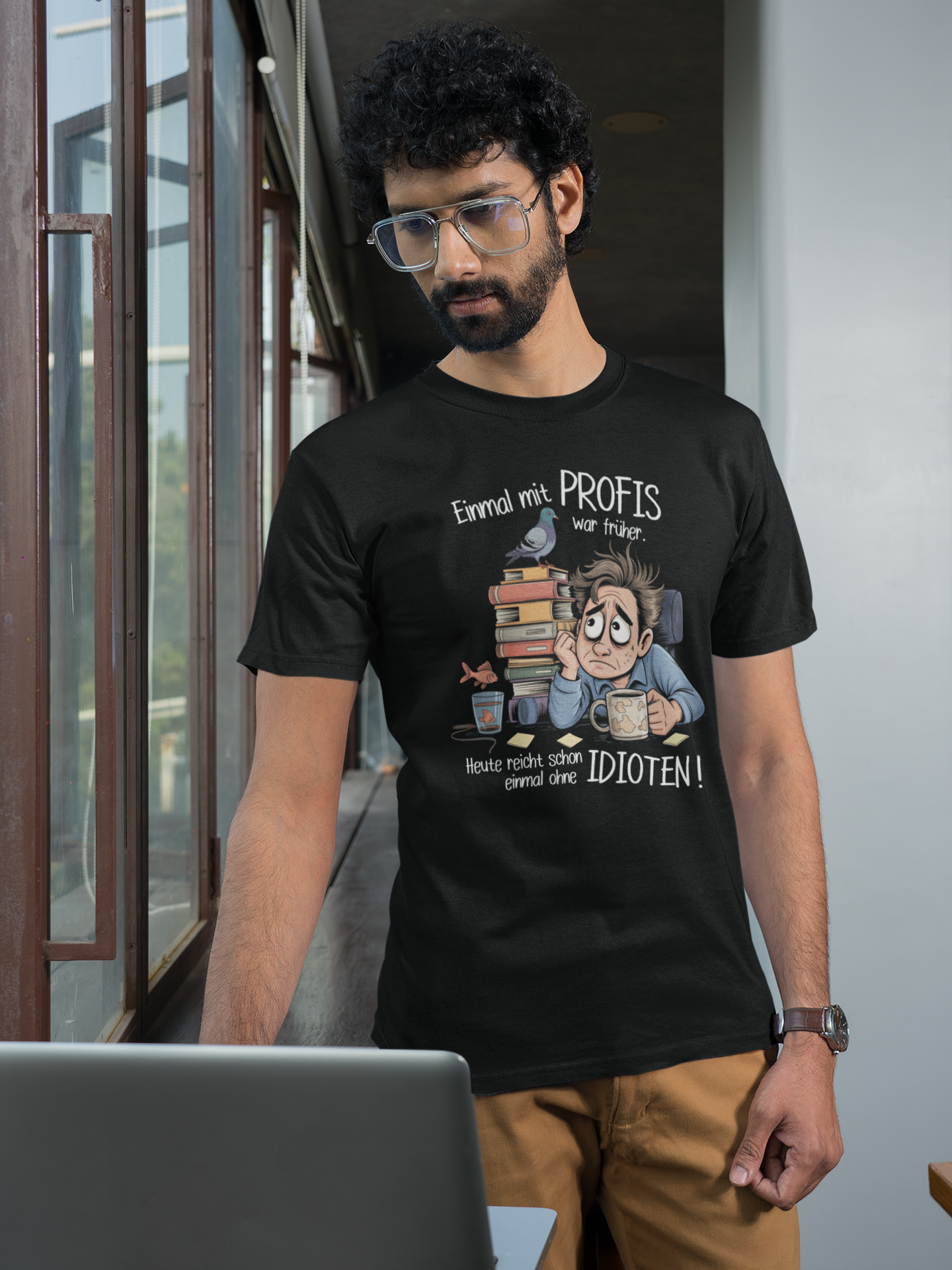 Junger Mann, stehend, mit Laptop, er trägt schwarzes Shirt mit dem Spruch „Einmal mit Profis war früher – Heute reicht schon ohne Idioten“ und Büro-Motiv. (Oberkörperaufnahme)