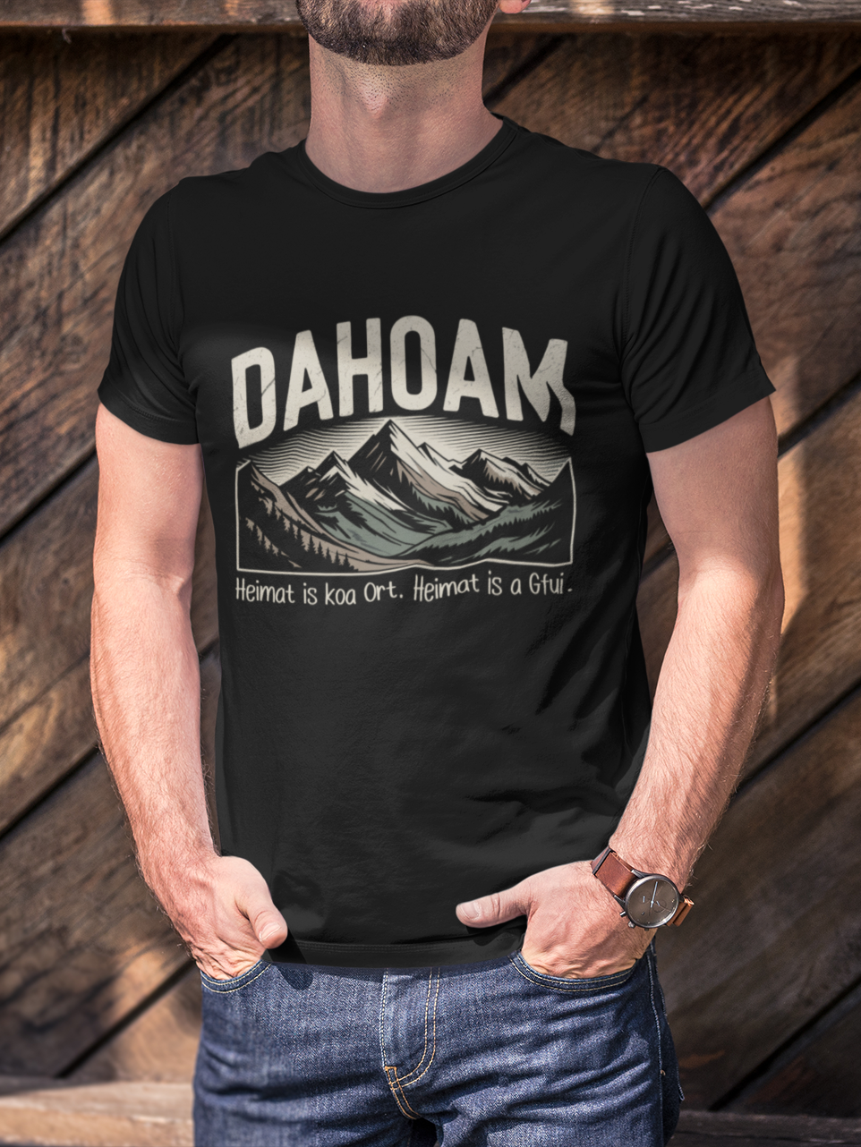 Mann trägt schwarzes T-Shirt mit Bergmotiv und Spruch „Dahoam – Heimat is koa Ort, Heimat is a Gfui“ (Oberkörper)